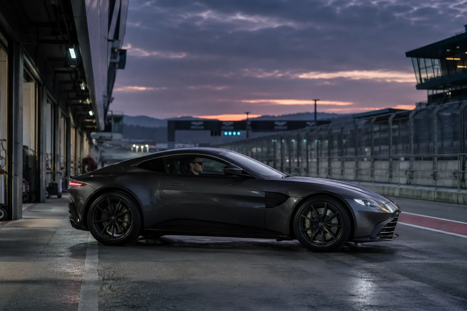 A graphite Aston Martin Vantage on a quiet pit-lane exit, contemporary executive driver inside